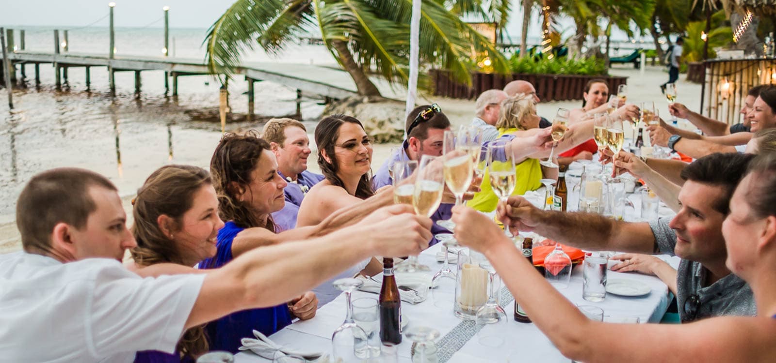 A group of people toasting with glasses of champagne at a festive outdoor table by the beach.