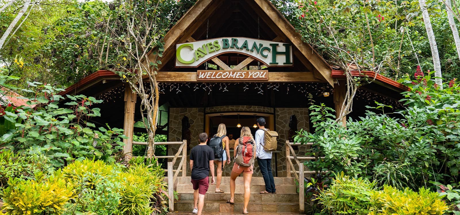 Four people walk toward the entrance of a rustic lodge surrounded by lush greenery. A wooden sign reads "Caves Branch Welcomes You," evoking a sense of adventure.