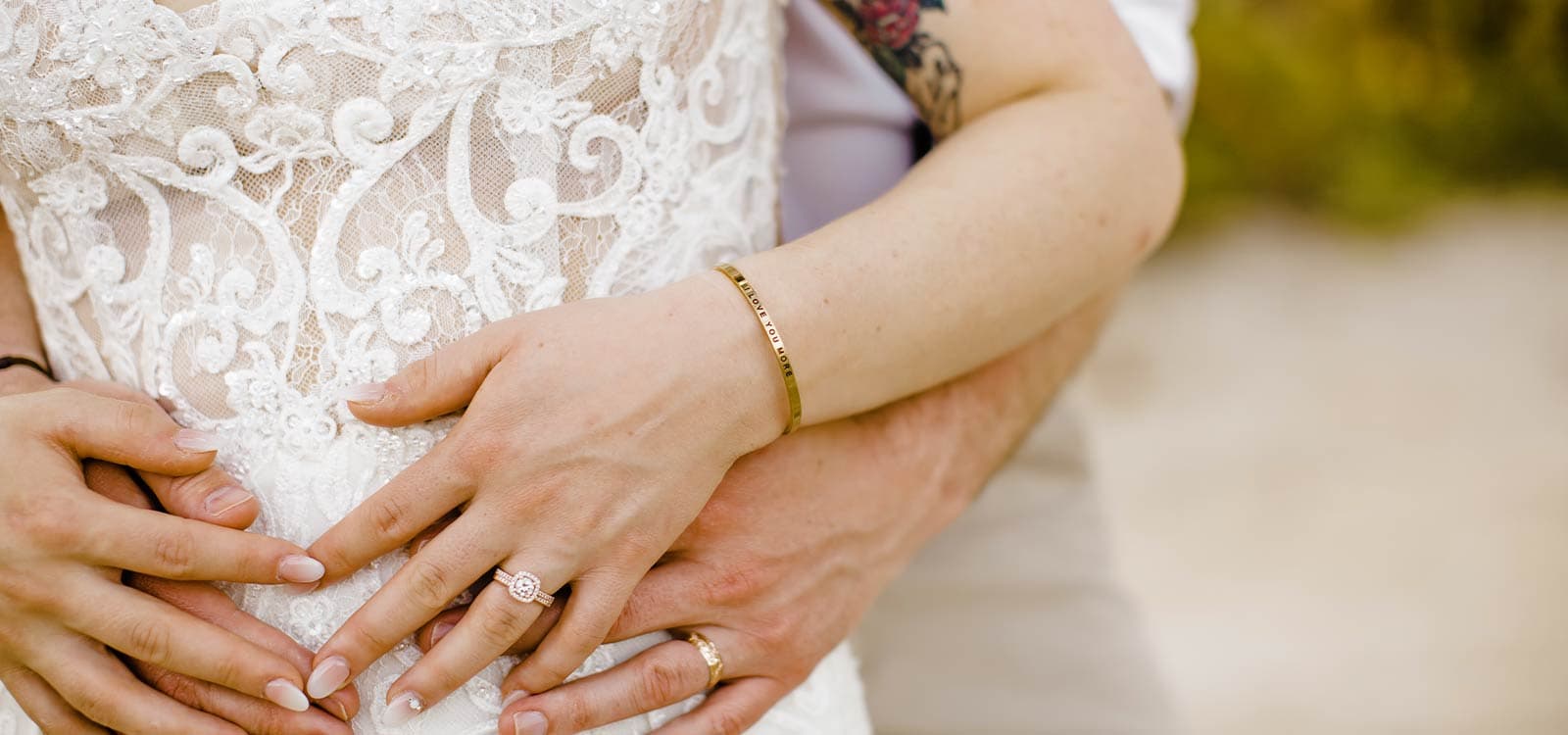 Close-up of a couple's embrace, showcasing intertwined hands over a lace wedding dress. Rings and a bracelet are visible, conveying love and intimacy.