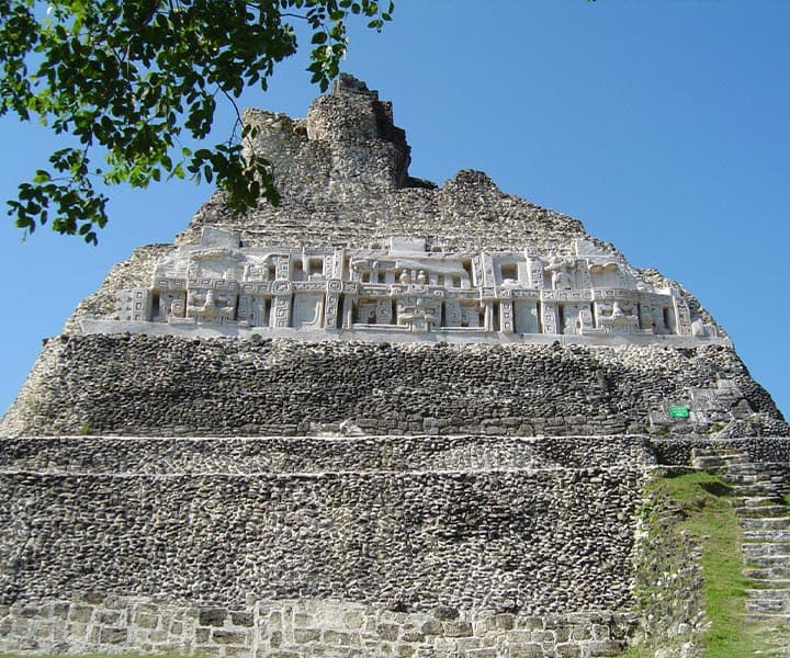 Ancient stone pyramid with intricate carvings surrounded by greenery under a blue sky.