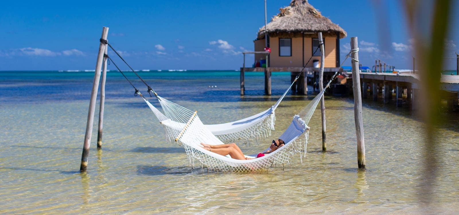 A woman relaxes on a white hammock above clear, shallow water, with a thatched-roof hut and blue sky in the background, exuding a tranquil, tropical vibe.